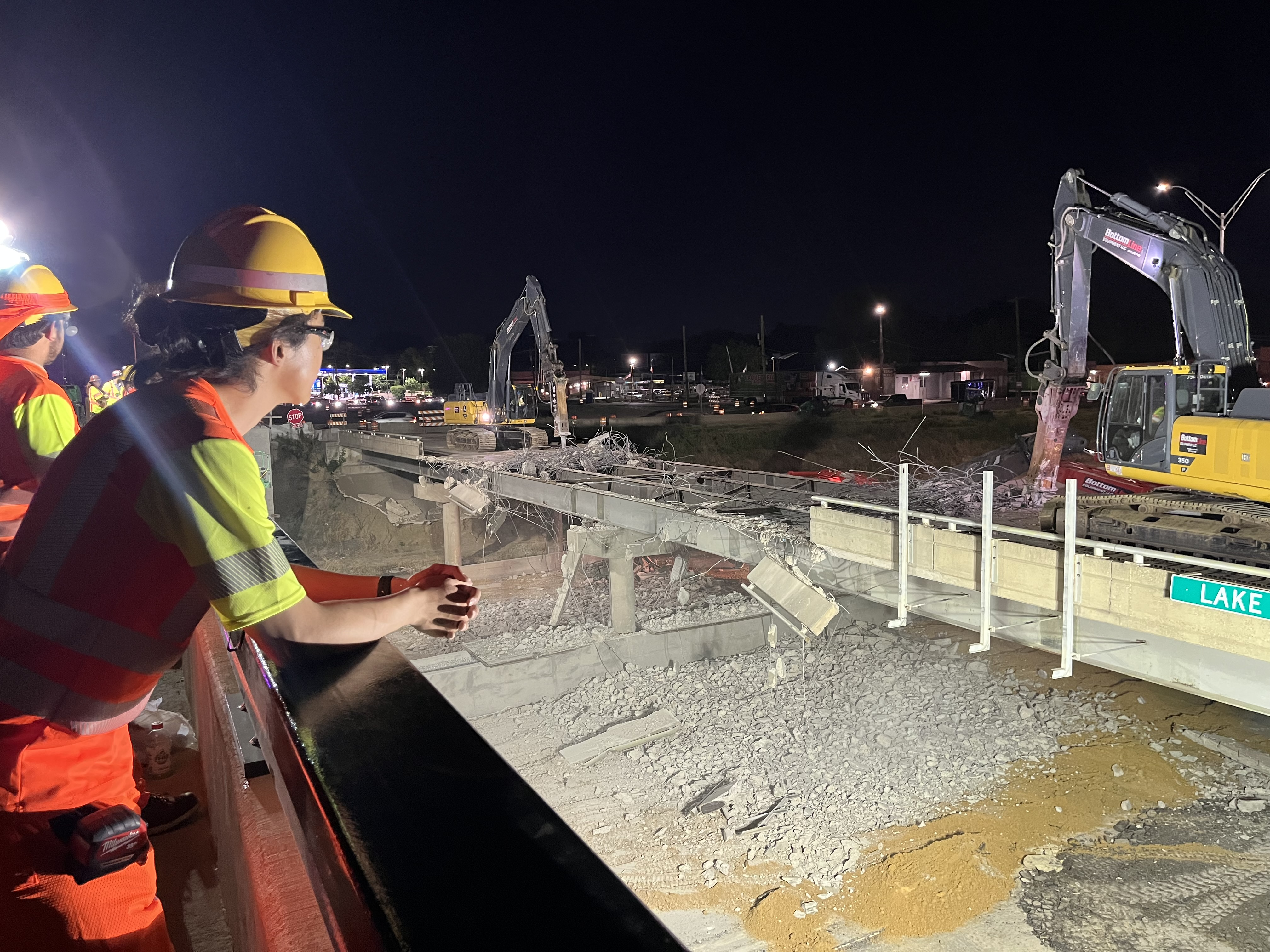 #4 Lake June bridge Dallas A worker in an orange vest and hard hat safety PPE is leaning on their elbows on the railing of one bridge looking over at the construction occuring across from them on the Lake June bridge.