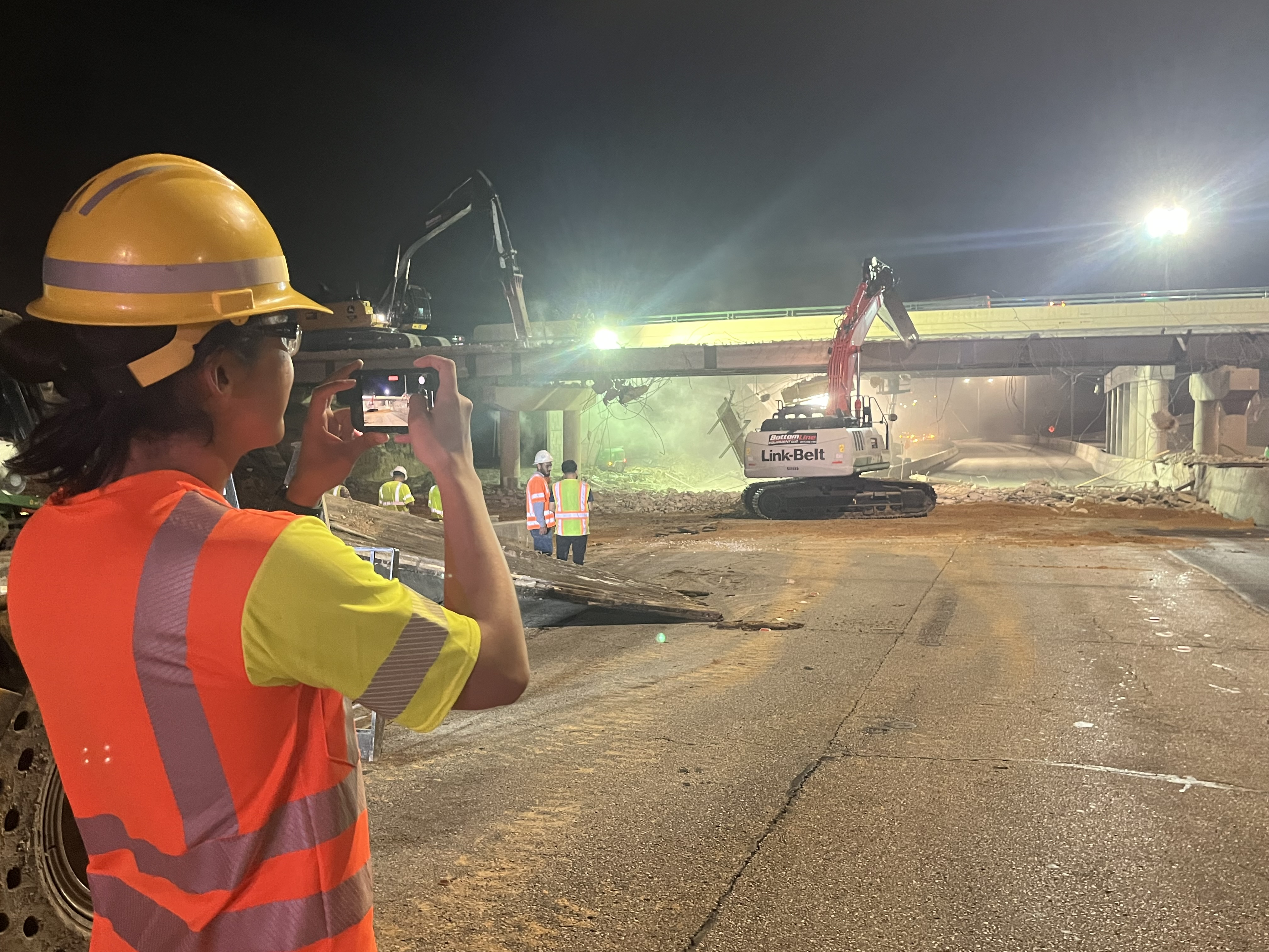 #3 Lake June bridge Dallas A worker in an orange PPE vest and hard hat is taking a picture of the construction site with his phone. In the construction site is the bridge, a link belt machine, and several other workers.