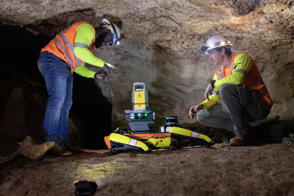 Cavern 3 A photo of two men in orange safety vests and hard hats crouched beside a four legged walking robot on a stone ledge inside Inner Space Cavern. One man is holding what appears to be a remote operating device.