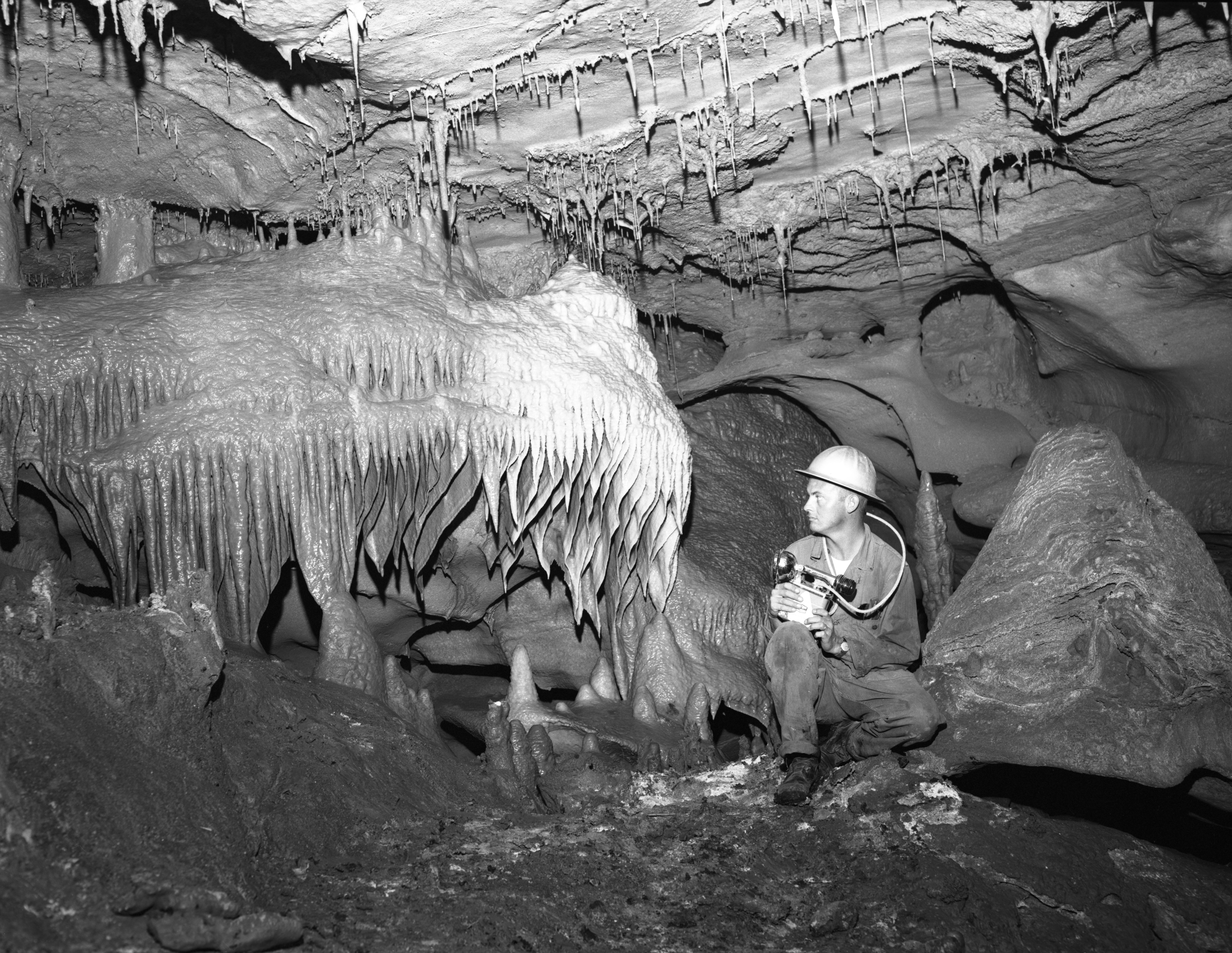 Cavern 5 An old black and white photo of a worker holding some sort of equipment and wearing a hard hat inside inner space cavern, crouched beside a strange rock formation that looks like it's melting.