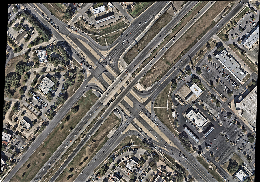 Aerial view of a displaced left-turn (DLT) intersection with an overpass, showing left-turn traffic rerouted across opposing lanes at signalized crossovers before the main intersection, enabling simultaneous left-turn and through movements.