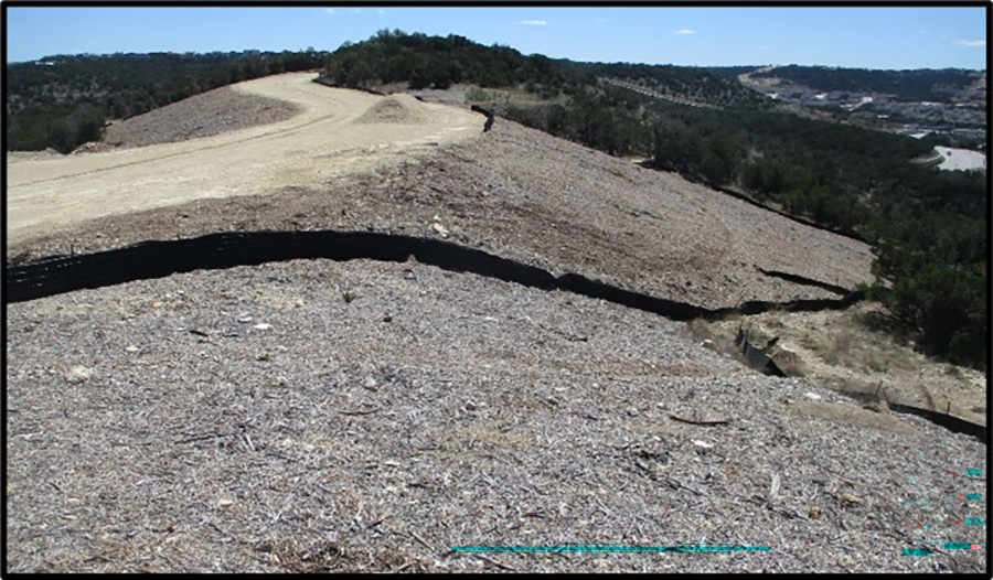 Una ladera cubierta con mantillo para estabilizar. También se ha instalado una valla de control de sedimentos a lo largo del contorno de la ladera. Hay un camino de tierra en la cima de la colina y hay vegetación escasa al fondo.