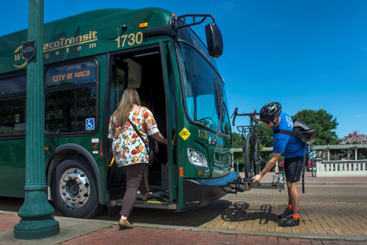 Una mujer con una camisa floreada sube a un gran autobús verde del sistema de transporte de Waco etiquetado con el número 1730 que está aparcado en una calle empedrada de Waco, Texas. Delante del autobús, un hombre con casco asegura una bicicleta al portaequipajes en la parte delantera del autobús. Es un día despejado y soleado con cielo azul asomando detrás del autobús.