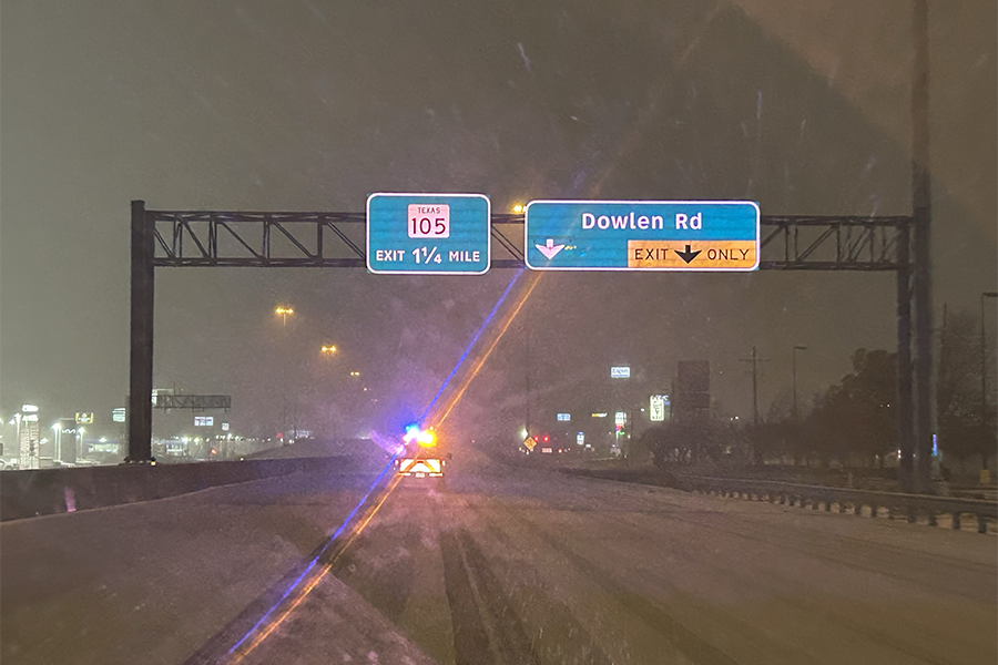Highway covered in snow with work truck ahead, lights on.