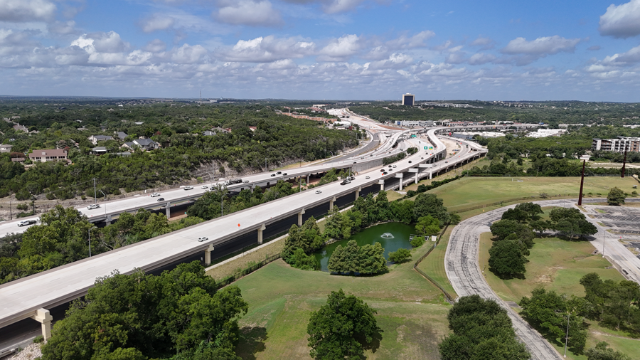 Image showing US 290 mainlane bridges traveling over the frontage roads, looking west. Trees are visible in the foreground and between the mainlane bridges, with the intersection with SH 71 in the distance. July 2026.