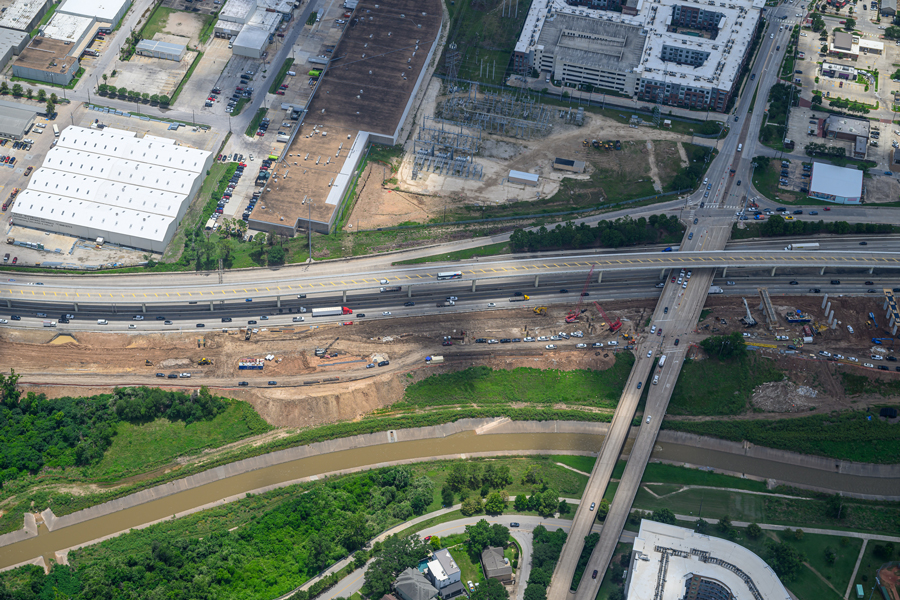 An aerial view of multiple roads intersecting with a long narrow dirt construction site running parallel to the west-east road. Many vehicles are present on both the road and in the construction site including a few cranes. There is also a canal full of water running parallel to the construction site. 